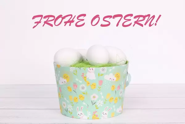 Eimer mit grünem Gras und weißen Ostereiern auf Holztisch mit Text Frohe Ostern auf weißem Hintergrund