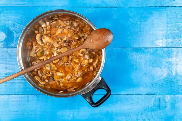 Ein Holzlöffel liegt auf einem Topf mit Hähnchen-Pilz Eintopf auf einem blauen Holztisch Draufsicht