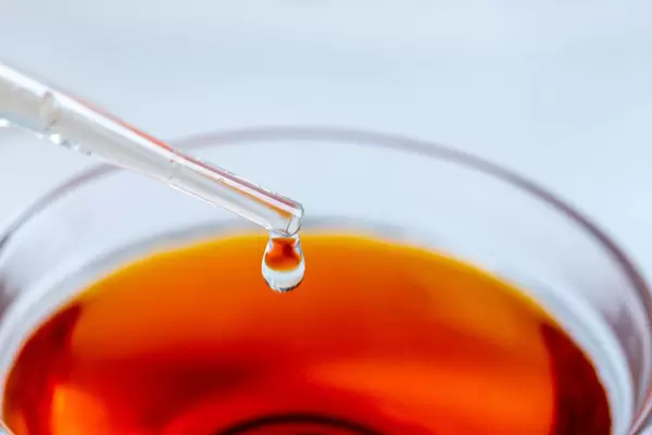 Ein Tropfen Flüssigkeit hängt an der Spitze einer Pipette über einer Schale mit roter Flüssigkeit Nahaufnahme