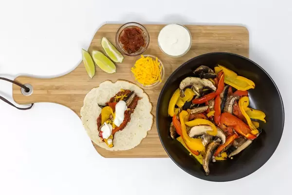 Eine Fajita, Limettenschnitte, scharfe Tomatensoße, Cheddar und Limettenschmand in kleinen Glasschälchen und Paprika mit Pilzen in einer schwarzen Schale auf einem Holzbrettchen auf weißem Hintergrund von oben fotografiert