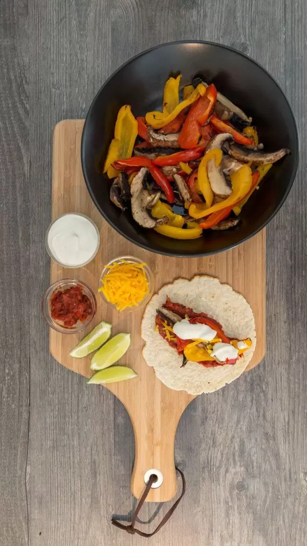 Eine Fajita, Limettenschnitte, scharfe Tomatensoße, Cheddar und Limettenschmand in kleinen Glasschälchen und Paprika mit Pilzen in einer schwarzen Schale auf einem Holzbrettchen