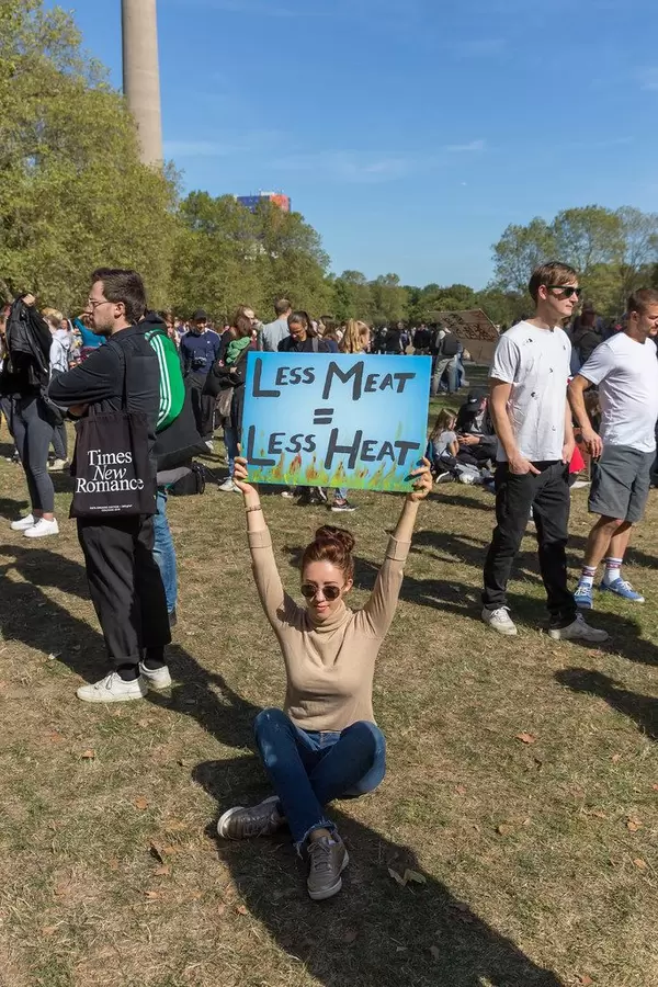 Eine Frau streckt das Plakat "Less Meat = Less Heat" in die Luft in einem Park