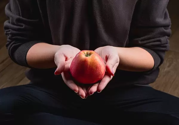 Eine in schwarz gekleidete Person im Schneidersitz hält einen frischen Apfel in beiden Händen
