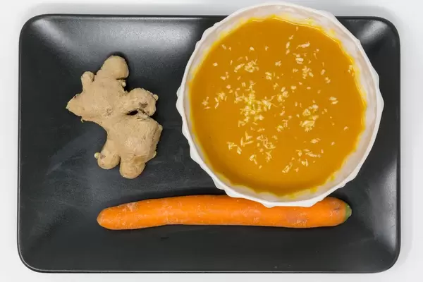 Eine Kürbis-Kokos-suppe mit Ingwer in einer Schüssel auf einem schwarzen Teller mit einer Karotte und einer Ingwerwurzel