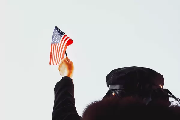 Eine Person in Schwarz angezogen hält eine amerikanische Fahne hoch vor weißem Hintergrund