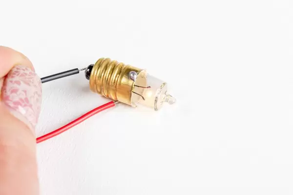 Eine Person schließt einen elektrischen Stromkreis mit schwarzem und rotem Stromkabel an einer kleinen Glühbirne vor weißem Hintergrund