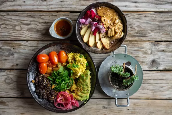 Eine Schüssel mit Porridge, frischen Früchten und Erdnussbutter und ein Teller mit Tofu, Avocado, Kartoffeln und schwarzen Bohnen von oben auf einem Holztisch fotografiert