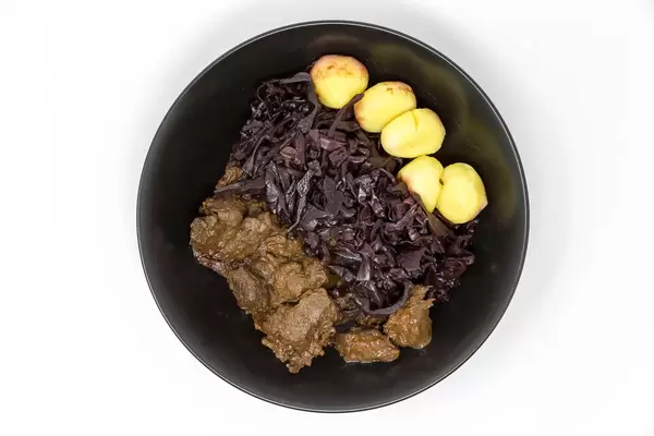 Eine Schüssel mit Rindergulasch nach traditioneller Art mit Apfelrotkohl und Miniknödeln in einer schwarzen Schüssel auf weißem Hintergrund in der Aufsicht