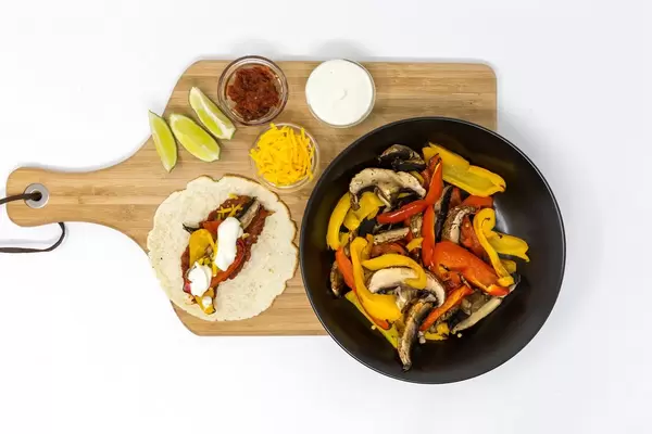 Eine vegetarische Fajita mit Paprika und Pilzen, scharfer Tomatensoße, Cheddar-Käse und Schmand mit Limettensaft mit dem Zutaten auf einem Brettchen auf weißem Hintergrund in der Aufsicht