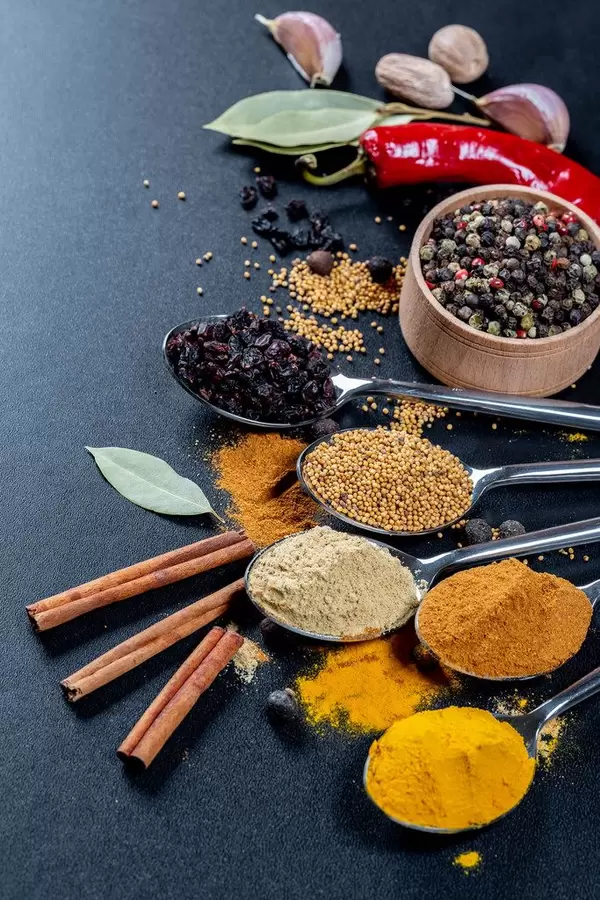 Elf Gewürze auf Löffeln auf schwarzem Hintergrund - Curry, Knoblauch und Kurkumapulver mit Senfkörnern und Berberitzen Zimtstangen, buntem Pfeffer, einer roten Chilischote, Lohrbeerblättern, Knoblauchzehen und Muskatnuss