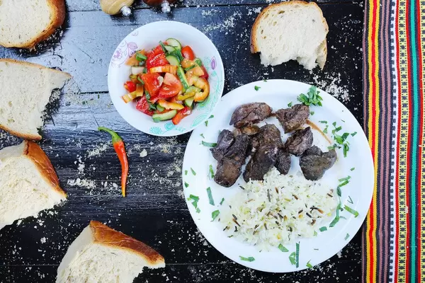 Entenleber mit Wildreis auf einem weißen Teller, mit einer Schüssel Gemüse und Weißbrot vor schwarzem Hintergrund