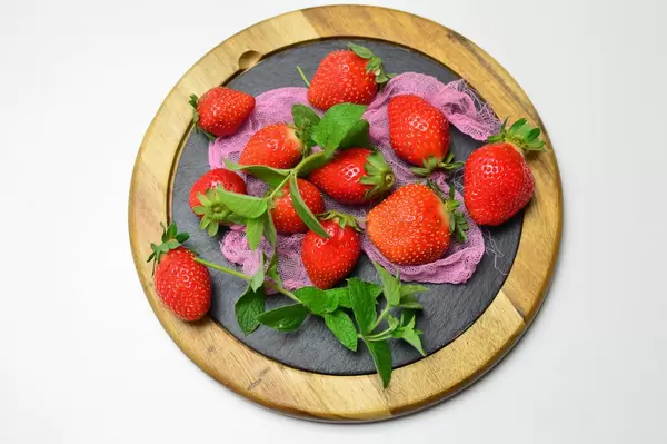 Erdbeeren mit Pfefferminzblättern auf runder Schieferplatte mit Holzrahmen und rosa Netz