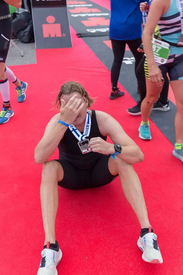 Erfolgreicher Ironman-Teilnehmer des 70.3-Triathlons sitzt erschöpft auf dem roten Teppich und hält seine Medaille hoch