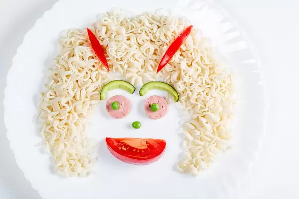 Face made of macaroni, sausage, avocado and tomato slice and green peas on white plate