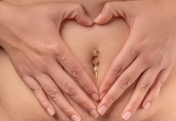 Female hands make a heart on the belly with piercing