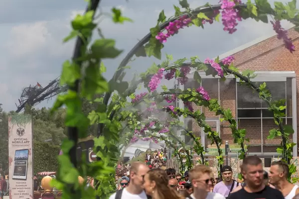Festivalbesucher kühlen sich unter Blumentoren mit Spritzwasser ab, neben ihnen ein Schild wirbt das offizielle Festival-App von Tomorrowland