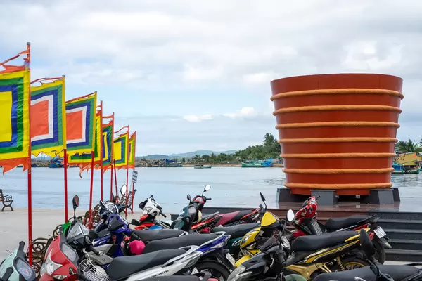 Fish Sauce Barrel Statue next to a Motorbike Parking and Buddhist Flags at Phu Quoc Harbour, Vietnam with Fishing Boats in the Background