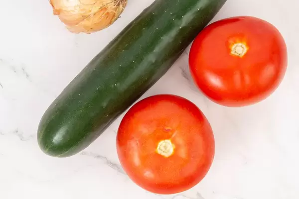 Flat-lay zeigt eine Salatgurke, gereifte Tomaten und eine Zwiebel, auf einer marmorierten Küchenarbeitsfläche