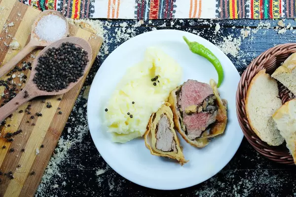 Flatlay auf schwarzem Hintergrund: gebackener Schweinenacken mit Kartoffelpüree und einem Jalapeño auf weißem Teller, serviert mit Weißbrot