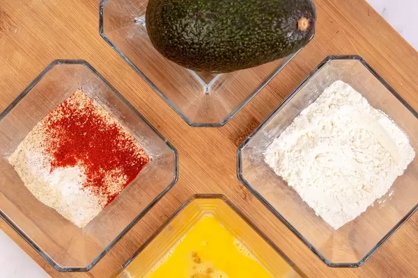 Flatlay mit ganzer Avocado, Mehl, Ei und Paniermehl mit scharfen Gewürzen in rechteckigen Glasschüsseln