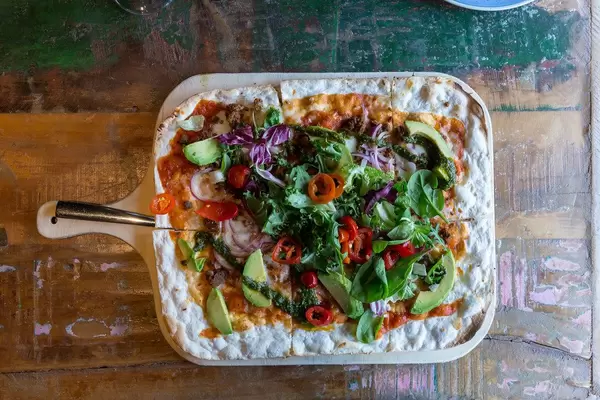 Flatlay shows flat bread on a colourful old wooden table, covered with pieces of Acovado, salad leaves, Chimichurri sauce and vegan meat substitute Härkis
