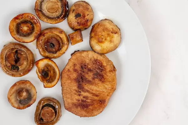 Flatlay zeigt braun-gebratenes Schweinefleisch neben zubereiteten Pilzen, auf einem weißen Teller