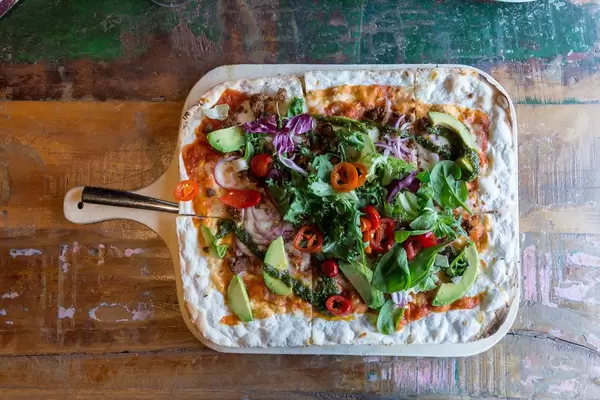 Flatlay zeigt Fladenbrot auf einem bunte Holztisch, belegt mit Acovadostückchen, Salatblättern, Chimichurri-Sauce und veganen Fleischersatz Härkis