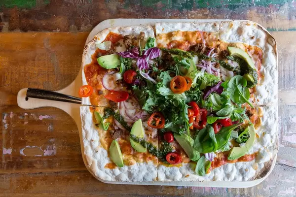Flatlay zeigt Fladenbrot auf einem Holzbrett, belegt mit Käse, Chipotle-Tomatensauce, Salat, Chimichurri, und veganem Chili-Härkis aus Saubohnen