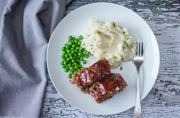 Flatlay zeigt zwei Scheiben vom Hackbraten, neben Erbsen und Kartoffelbrei, auf einem Vintagetisch