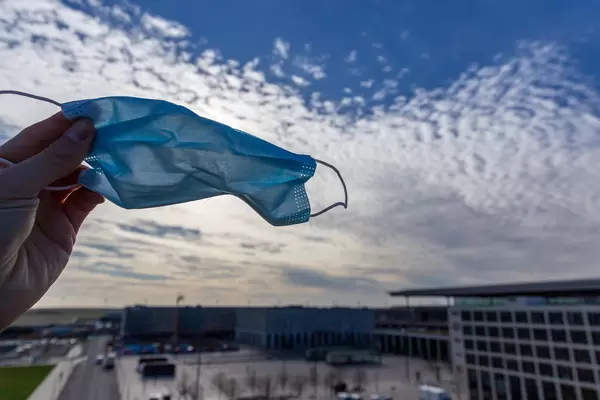 Flughafen BER: Hand hält eine OP Maske mit dem Himmel im Hintergrund und dem ²BAC Gebäude