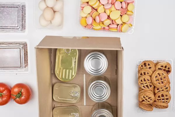 Food bank: top view of tin cans in a cardboard box surrounded by tomatoes, eggs, cookies, dried fruit