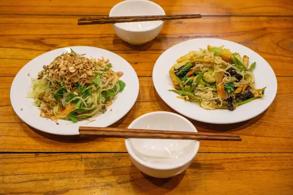 Food Photo of a Plate of Papaya Salad with Carrots, Mint, Fried Onions, Peanuts and Fishsauce next to a Plate of Fried Noodles with Vegetables on a Wooden Table with Bowls and Chopsticks