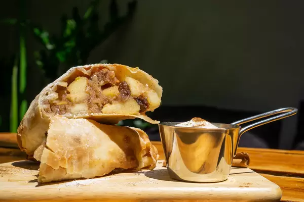 Food Photo of Apple Strudel Desssert with Raisin and Fresh Apples on a Wooden Board with Whipped Cream and Cinnamon in a Restaurant