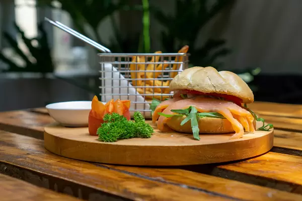 Food Photo of Smoked Salmon Sandwich with Arugula, Tomato and Honey Mustard Sauce in a German Bread Roll on a Wooden Board with Parsley, Tomatoes and a Basket with French Fries in a Restaurant