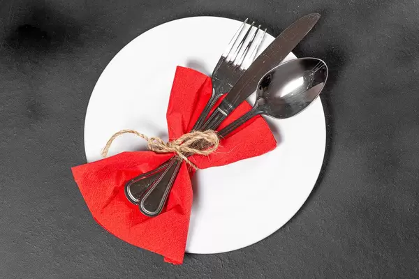 Fork, knife and spoon with red napkin and white plate on black background. Top view (Flip 2019)