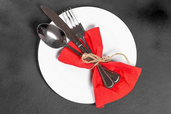 Fork, knife and spoon with red napkin and white plate on black background. Top view