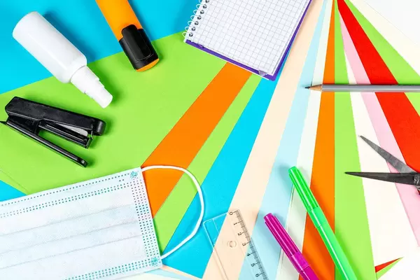 Frame with school supplies and medical mask on colored paper background, top view