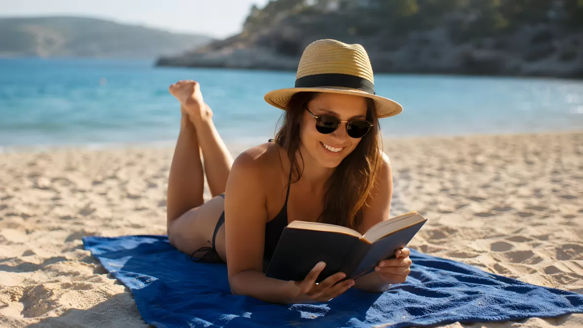 Frau liest Buch am Strand mit Strohhut und Sonnenbrille