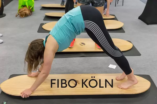 Frau macht Yoga- und Fitnessübungen auf einem Holzbrett, über dem Bildtitel "Fibo Köln"