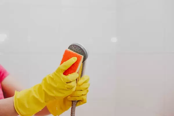 Frau mit gelben Gummihandschuhen und einem Schwamm reinigt die Duschbrause im Badezimmer am Putztag