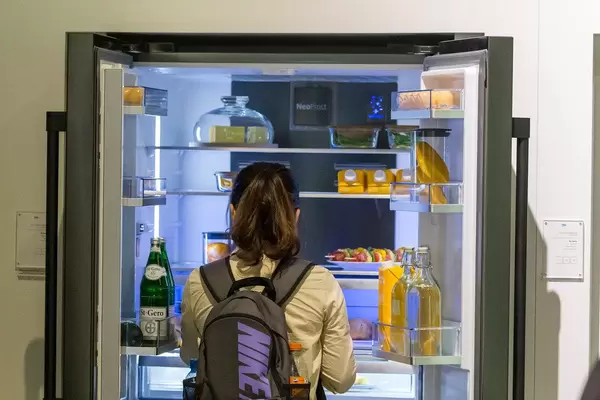 Frau mit Rucksack steht vor einem geöffneten Doppeltür-Kühlschrank und blickt auf gesunde Lebensmittel
