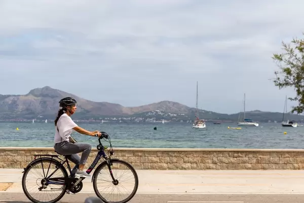 Frau mit weißer Bluse und schwarzem Fahrradhelm fährt Fahrrad am Meer entlang auf Mallorca