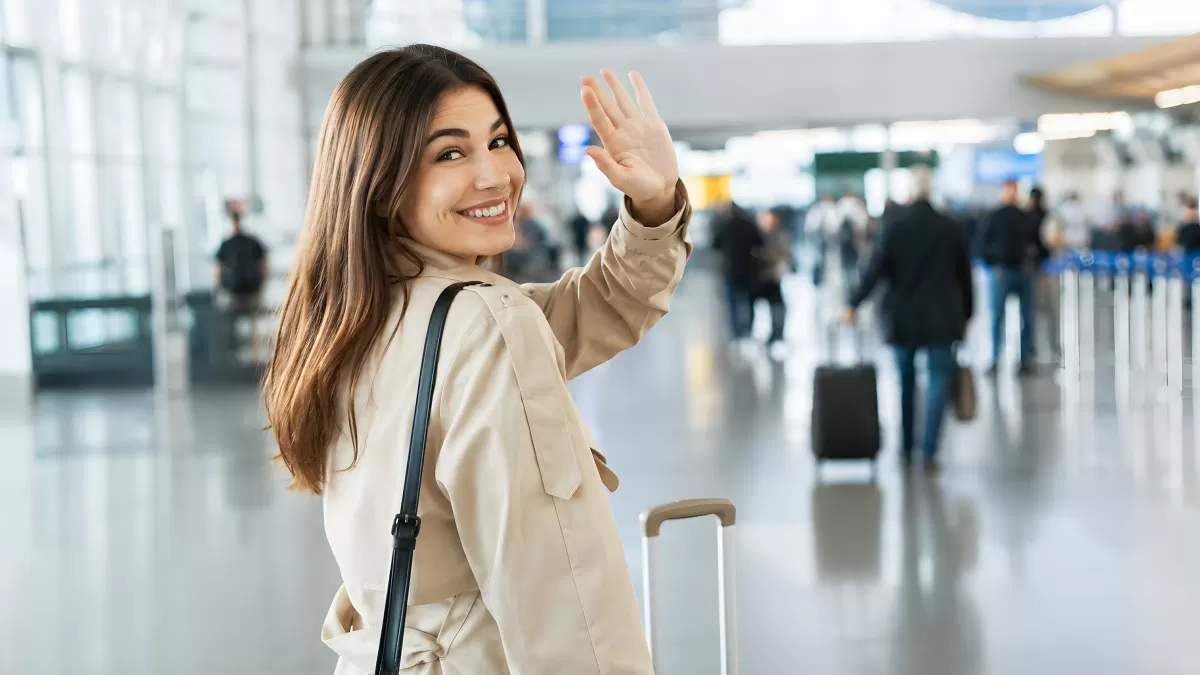 Frau winkt lächelnd am Flughafen mit Trolley