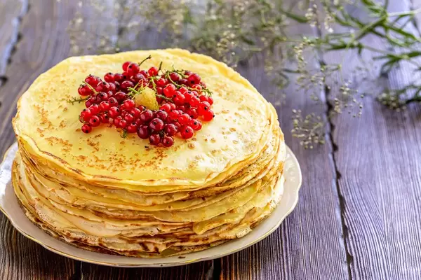 Fresh thin pancakes with red currant berries on a brown wooden background with belima flowers