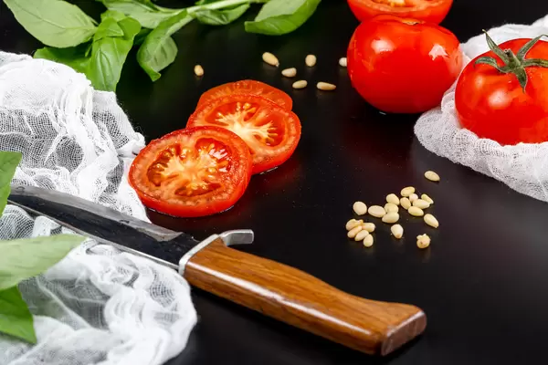 Fresh tomatoes with pine nuts, basil, knife and gauze on black background