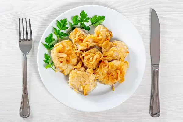 Fried heck fish with onion and fresh parsley on white table with knife and fork