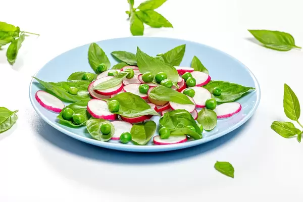 Frische Basilikumblätter, Radieschen und grüne Erbsen auf blauem Teller mit weißem Hintergrund
