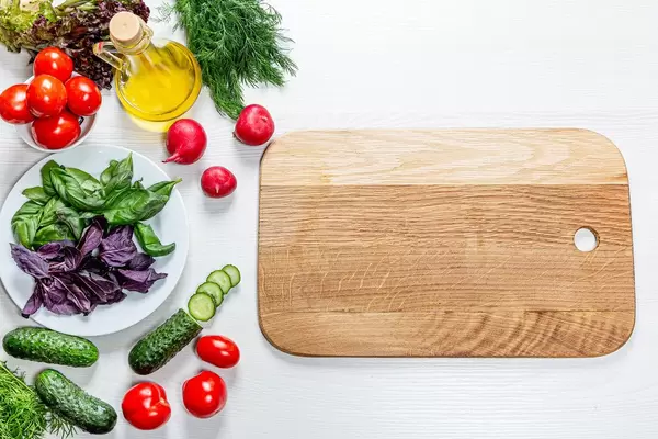 Frische Tomaten, Gurken, Radieschen, Basilikum und Zitrone neben einem Holzbrett auf einem weißen Tisch top view