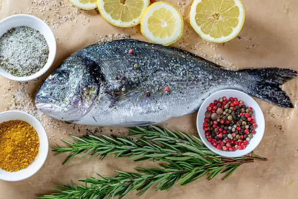 Frischer Goldbrasse-Fisch / Doradefisch, mit Rosmarinstengel, Pfefferkörnern und Meeressalz, auf einem Backpapier, aus der Sicht von oben