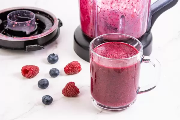 Frischer Smoothie mit Blaubeeren und Himbeeren in Glas vor benutztem Saftmixer